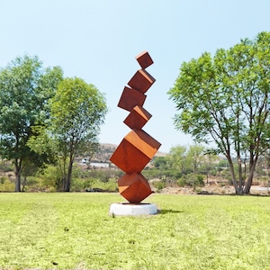 K&ouml;nnte beinhalten: Eine moderne Au&szlig;enskulptur aus gestapelten, rostorangefarbenen Metallw&uuml;rfeln. Die Skulptur steht auf einem wei&szlig;en Sockel und erhebt sich vor dem Hintergrund von gr&uuml;nem Gras, B&auml;umen und einem klaren blauen Himmel. Das Kunstwerk ist eine abstrakte Darstellung des Gleichgewichts.