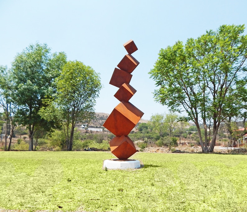 K&ouml;nnte beinhalten: Eine moderne Au&szlig;enskulptur aus gestapelten, rostorangefarbenen Metallw&uuml;rfeln. Die Skulptur steht auf einem wei&szlig;en Sockel und erhebt sich vor dem Hintergrund von gr&uuml;nem Gras, B&auml;umen und einem klaren blauen Himmel. Das Kunstwerk ist eine abstrakte Darstellung des Gleichgewichts.