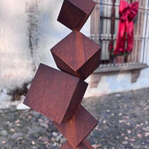 K&ouml;nnte beinhalten: Eine moderne, abstrakte Skulptur aus gestapelten, geometrischen W&uuml;rfeln. Die Skulptur besteht aus verwittertem, r&ouml;tlich-braunem Metall mit einer strukturierten Oberfl&auml;che. Die W&uuml;rfel sind in scheinbar prek&auml;rer Balance angeordnet.