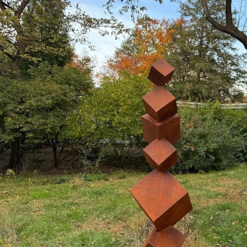 K&ouml;nnte beinhalten: Eine hohe Au&szlig;enskulptur aus gestapelten, rostbraunen Metallw&uuml;rfeln. Die W&uuml;rfel sind in einer vertikalen S&auml;ule angeordnet, wobei jeder W&uuml;rfel leicht versetzt zum darunter liegenden ist. Die Skulptur steht vor einem Hintergrund aus gr&uuml;nen B&auml;umen und Gras.