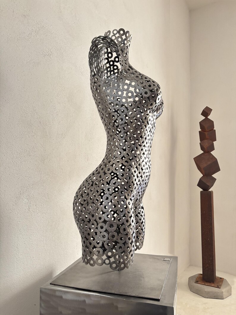 May include: A metal sculpture of a female torso constructed from numerous circular metal pieces. The sculpture is displayed on a square metal base. A second sculpture of stacked cubes on a metal pole is in the background.