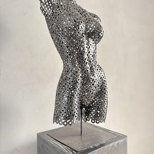 May include: A metal sculpture of a female torso, constructed from numerous small, circular metal pieces. The sculpture is mounted on a square, gray metal pedestal. The artwork is displayed against a neutral background, highlighting the form and texture.