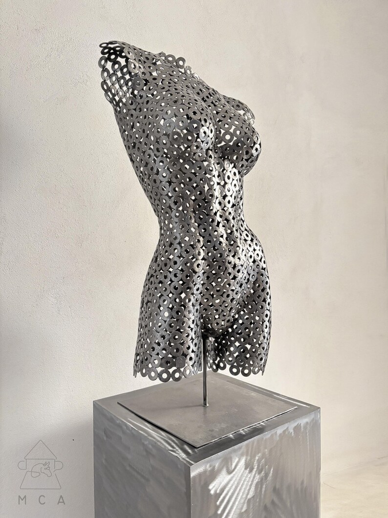 May include: A metal sculpture of a female torso, constructed from numerous small, circular metal pieces. The sculpture is mounted on a square, gray metal pedestal. The artwork is displayed against a neutral background, highlighting the form and texture.
