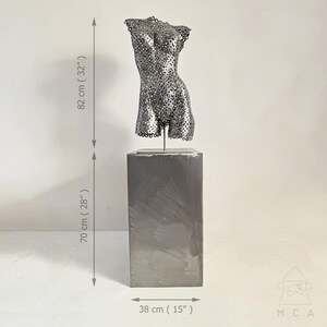 May include: A metal sculpture of a torso, 32 inches tall, with a perforated design, mounted on a 28-inch tall rectangular pedestal. The pedestal is 15 inches wide. The sculpture is a modern art piece.