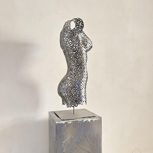May include: A silver metal sculpture of a torso with a perforated design, mounted on a square, brushed metal pedestal. The sculpture is a stylized representation of the human form, with a focus on the curves of the body.