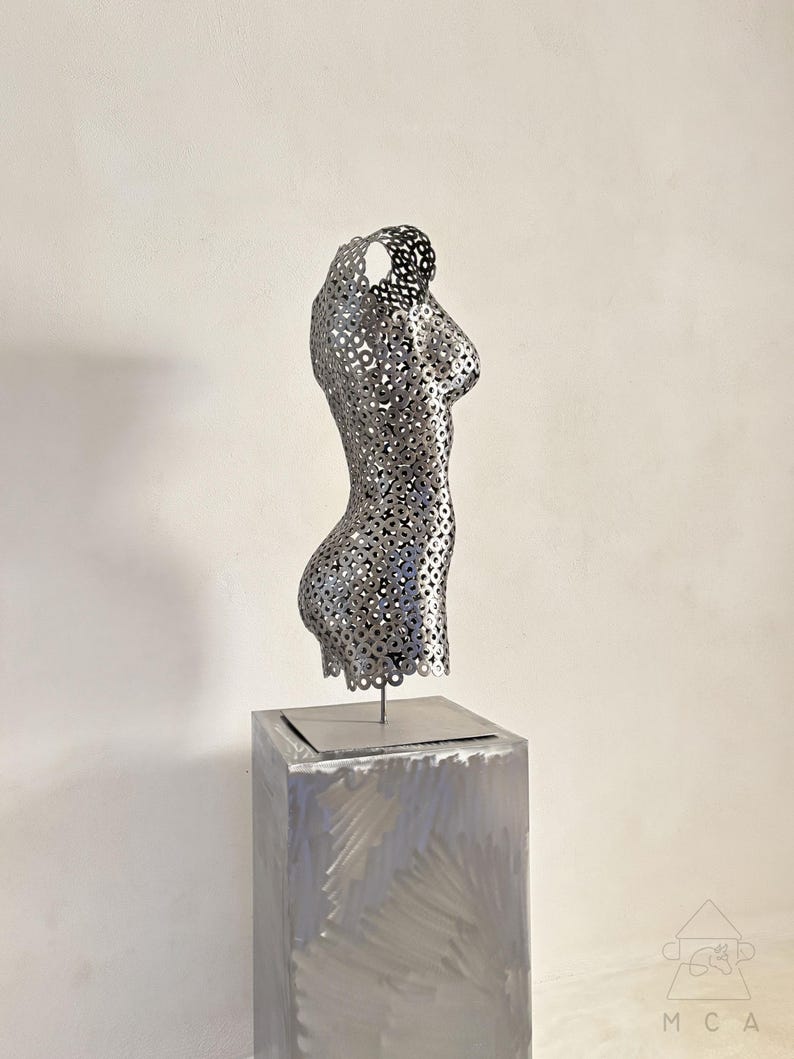 May include: A silver metal sculpture of a torso with a perforated design, mounted on a square, brushed metal pedestal. The sculpture is a stylized representation of the human form, with a focus on the curves of the body.