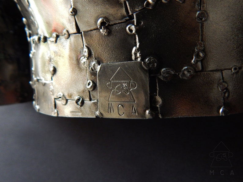 Może przedstawiać: Zbliżenie metalowej rzeźby o geometrycznym wzorze. Rzeźba wykonana jest z wielu małych kawałk&oacute;w metalu, kt&oacute;re są ze sobą spawane. Rzeźba jest podpisana "MCA" u dołu.