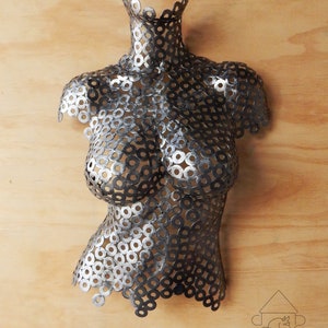 May include: A silver metal sculpture of a torso made from many small metal rings. The sculpture is mounted on a wooden surface.
