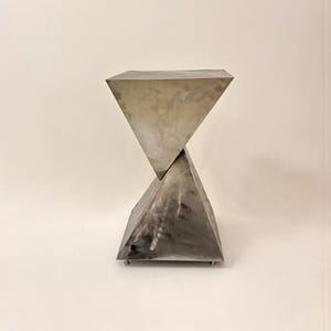 Puede incluir: Una mesa auxiliar moderna y geométrica hecha de metal cepillado. La mesa presenta dos formas piramidales que se cruzan, creando un diseño único en forma de reloj de arena. El metal tiene un acabado plateado y está soportado por pequeños pies.