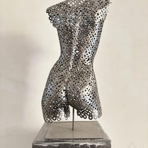 May include: A metal sculpture of a torso, constructed from numerous circular metal pieces. The sculpture is mounted on a square metal base and stands against a plain white background. The sculpture's form is detailed and textured, with a focus on the back and shoulders.