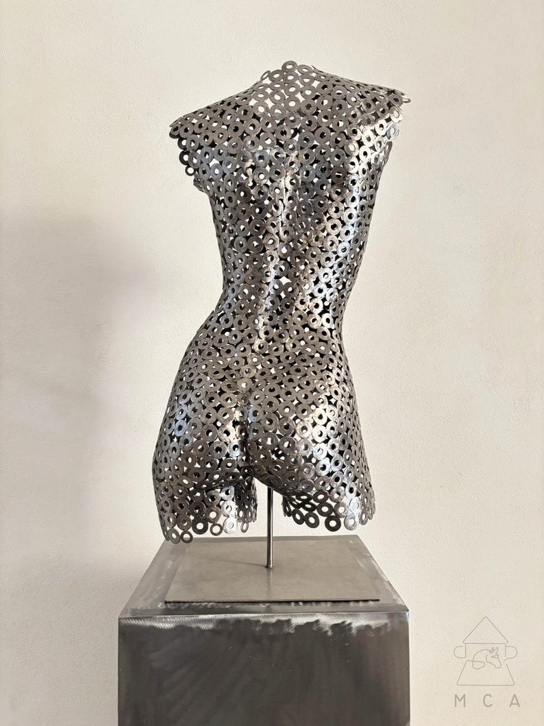 May include: A metal sculpture of a torso, constructed from numerous circular metal pieces. The sculpture is mounted on a square metal base and stands against a plain white background. The sculpture's form is detailed and textured, with a focus on the back and shoulders.