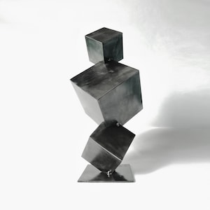 Könnte beinhalten: Eine moderne Skulptur aus drei übereinander gestapelten Metallwürfeln. Die Würfel sind alle gleich groß und aus dunkelgrauem Metall gefertigt. Die Skulptur steht auf einem kleinen Metallsockel.