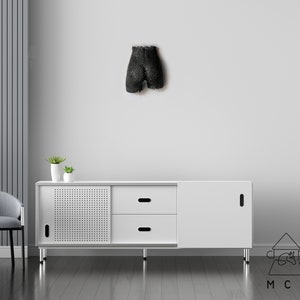 May include: A white cabinet with two drawers and a door. The cabinet has a black metal frame and legs. The cabinet is sitting on a wooden floor. There is a black sculpture on the wall behind the cabinet.