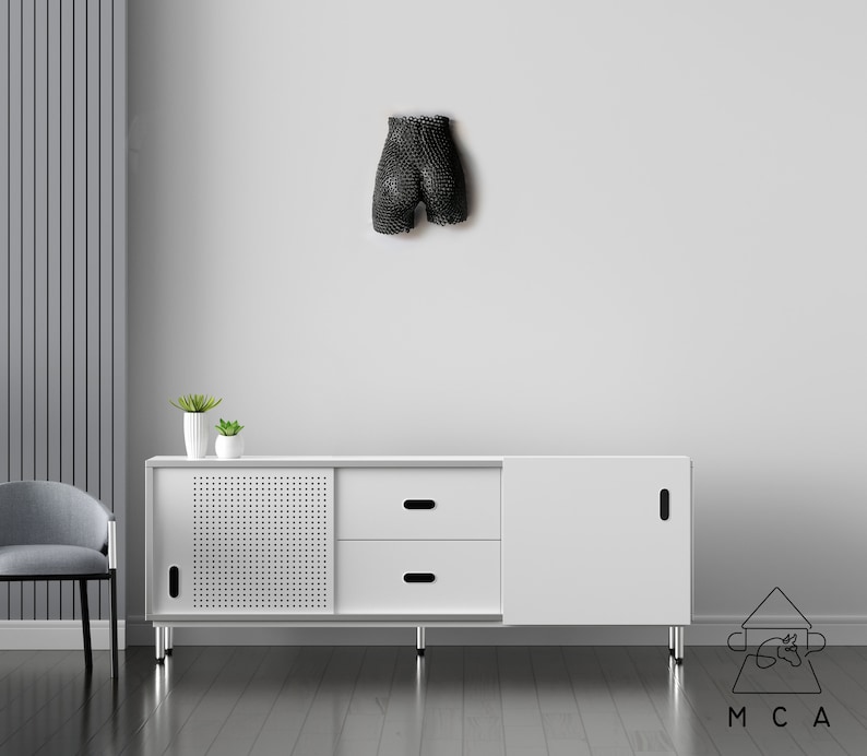 May include: A white cabinet with two drawers and a door. The cabinet has a black metal frame and legs. The cabinet is sitting on a wooden floor. There is a black sculpture on the wall behind the cabinet.