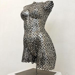 May include: A metal sculpture of a female torso, constructed from numerous small, circular metal pieces. The sculpture is mounted on a metal stand and base, showcasing the form's curves and details. The artwork is displayed against a white background.