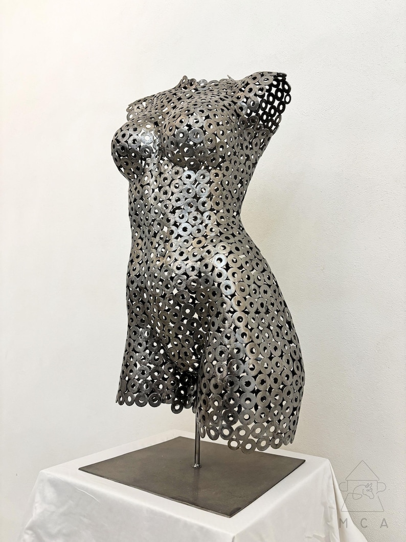 May include: A metal sculpture of a female torso, constructed from numerous small, circular metal pieces. The sculpture is mounted on a metal stand and base, showcasing the form's curves and details. The artwork is displayed against a white background.