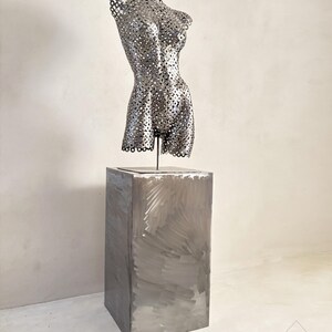 May include: A metal sculpture of a torso with a perforated design, mounted on a rectangular metal pedestal. The sculpture is silver-toned and the pedestal has visible brushstrokes. The overall aesthetic is modern and artistic.