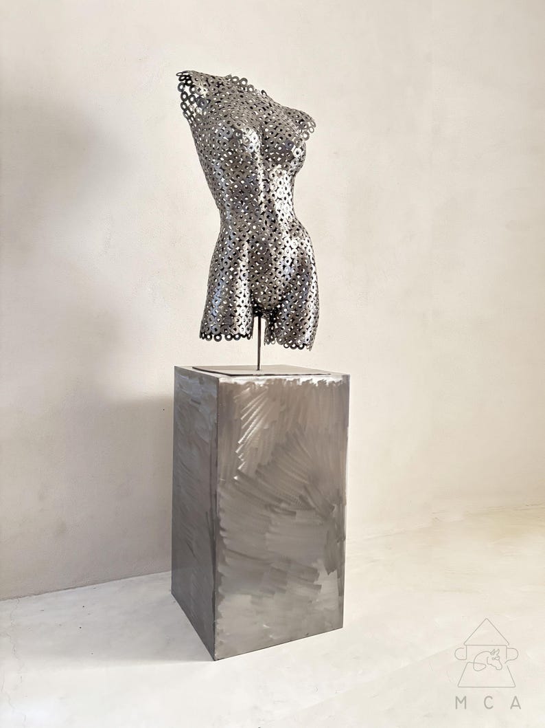 May include: A metal sculpture of a torso with a perforated design, mounted on a rectangular metal pedestal. The sculpture is silver-toned and the pedestal has visible brushstrokes. The overall aesthetic is modern and artistic.