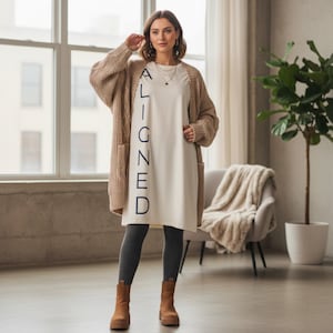 Puede incluir: Una mujer con una rebeca beige sobre un vestido color crema con la palabra "ALIGNED" impresa verticalmente en negro. También lleva leggings grises y botas marrones. Una silla con una manta y una planta en maceta están en el fondo.