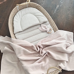May include: A woven wicker baby bassinet with a gingham patterned cushion and a soft pink blanket. The bassinet includes a wooden ring toy with a fabric bunny ear attachment. The blanket is draped over the side of the bassinet.
