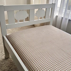 Puede incluir: Cama de madera blanca con cabecero con dos recortes de ovejas. La cama tiene un colchón cubierto con un patrón de cuadros marrones y blancos. La cama está sobre una alfombra de color claro.