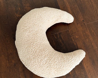 Op de afbeelding: Een crèmekleurig, halvemaanvormig kussen gemaakt van zacht, pluche stof. Het kussen ligt op een donkerbruine houten vloer.
