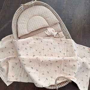 Op de afbeelding: Een geweven rieten wieg met een beige en witte geruite voering en een beige mousseline deken met een paardenbloemprint.