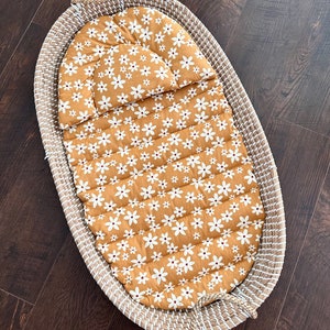 May include: An oval woven basket with a mustard-coloured, quilted insert. The insert features a pattern of white daisy flowers. The basket has rope handles and is designed for a baby or infant.