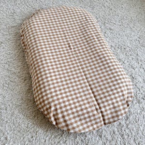 May include: A brown and white gingham patterned pillow. The pillow is oblong and has rounded edges. The fabric has a classic checkered design, with alternating squares of brown and white. The pillow is placed on a light gray, textured surface.