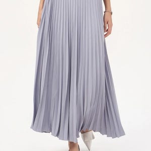 May include: A long, pleated skirt in a soft lavender colour. The skirt has a flowing silhouette and is paired with white shoes. The pleats create a vertical texture, adding visual interest to the garment. The skirt is part of a fashion ensemble.