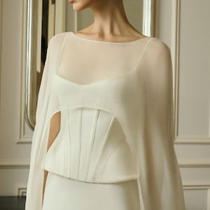 May include: A floor-length white gown featuring a structured corset bodice with vertical seams and a sheer, lightweight cape overlay that drapes over the shoulders. The garment has a wide, rounded neckline and a clean, solid white finish throughout.