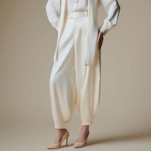 May include: An off-white long-sleeved button-down blouse with a matching attached scarf collar and high-waisted, loose-fitting tapered trousers. The ensemble is crafted from a smooth, solid-colored fabric and is paired with pointed-toe beige heels.