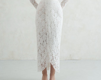 May include: A white lace midi dress featuring long sleeves, a square neckline, and a structured bodice. The garment includes a delicate fabric flower accent on the left shoulder and a wrap-style skirt with a high-low hemline, crafted from floral lace.