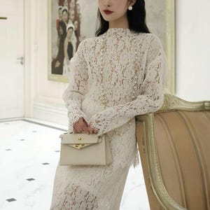May include: A woman in a white lace dress and matching long-sleeved top, holding a small beige handbag. The outfit features a high neckline and pearl accents. She is standing near a striped chair in a well-lit room.