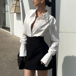 Puede incluir: Un conjunto femenino con una camisa blanca abotonada con puños anchos y una minifalda negra. La camisa está desabrochada en la parte superior. Un pequeño bolso negro cuelga del hombro. Pendientes dorados.