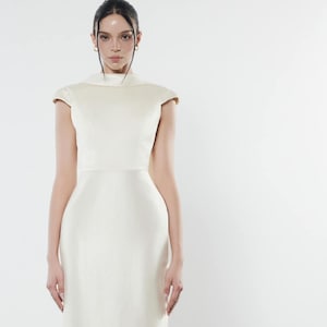 Puede incluir: Un vestido maxi blanco con cuello alto y mangas cortas. El vestido tiene un corpiño ajustado y una falda fluida.