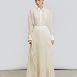 May include: A cream-coloured, pleated skirt and long-sleeved blouse ensemble. The blouse has a collared neckline and button-down front. The skirt is floor-length. The outfit is made of a lightweight, flowing fabric.