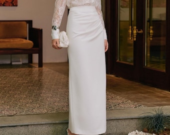May include: A long-sleeved white lace button-down shirt with a pointed collar paired with a floor-length white maxi skirt featuring a side-ruched detail. The outfit is completed with a small white clutch bag and pointed-toe white heels.