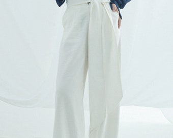 May include: Off-white wide-leg pants with a high waist and a wrap-around fabric detail. The pants have a relaxed fit and are paired with neutral-toned sandals. The model is wearing a dark blue shirt. The pants are made of a light-colored fabric.