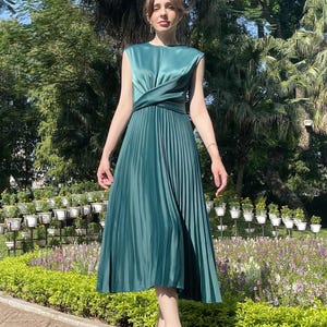 Peut inclure: Une femme portant une robe midi pliss&eacute;e verte avec un corsage crois&eacute; et une finition satin&eacute;e. Elle se tient dans un jardin avec de la verdure et des fleurs.