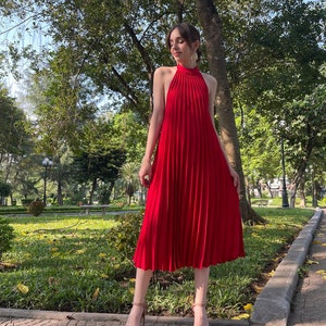Pode incluir: Uma mulher vestindo um vestido vermelho de al&ccedil;as com pregas. O vestido &eacute; midi e sem mangas. Ela est&aacute; em p&eacute; em um caminho de paralelep&iacute;pedos em um parque.