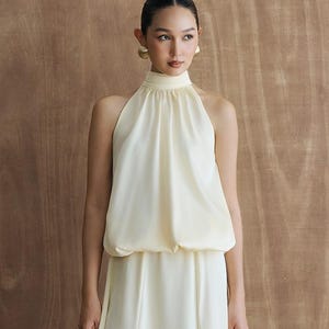 May include: A floor-length, pale yellow sleeveless dress featuring a high halter neckline with a gathered bodice that creates a blouson effect at the waist, transitioning into a smooth, flowing skirt that reaches down to the ankles.