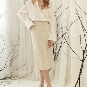 Puede incluir: Una mujer con una camisa de lino blanca con botones negros y una falda midi beige abotonada. Lleva sandalias de tiras rosa claro. El fondo presenta cortinas blancas y ramas decorativas.