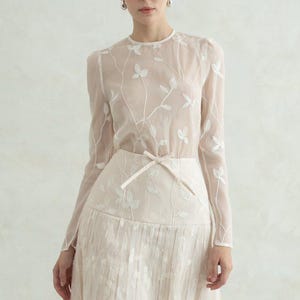 May include: A long-sleeved, floor-length dress in a pale cream hue. The bodice is made of sheer fabric with white embroidered leaf patterns. The skirt features a pleated design with a thin ribbon tie at the waist and matching white leaf embroidery.