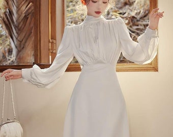 May include: A white long-sleeved dress with a high neck and a fitted bodice. The dress has a flowing skirt and is made of a lightweight fabric. The model is holding a white clutch purse with a pearl chain.