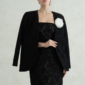 Op de afbeelding: Een zwarte midi-jurk met een vierkante halslijn en een bijpassende zwarte blazer die over de schouders is gedrapeerd. Het ensemble heeft een tonaal bloemenpatroon in de stof. Een grote witte stoffen bloem is op de revers gespeld.