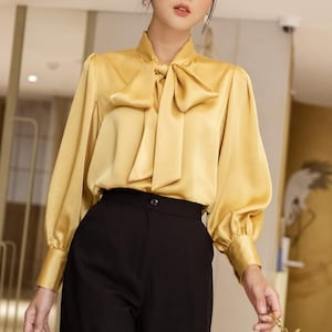 May include: A golden yellow satin blouse with a large bow at the neck and long sleeves, paired with black trousers. A small black purse with a gold chain is held in the hand. The outfit is suitable for formal or business attire.