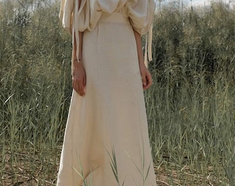 May include: A woman wearing a long, cream-colored linen dress with a gathered waist and cinched sleeves. The dress has a relaxed fit and a flowing skirt. She is standing in a field of tall grass.