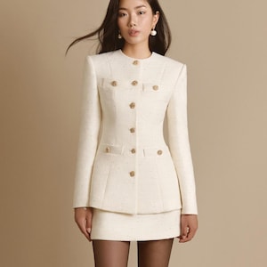 May include: A cream-colored tweed jacket and matching mini skirt set. The jacket features a round neckline, long sleeves, and a front button closure with gold-toned buttons. Two small flap pockets are positioned on the chest and two on the waist.