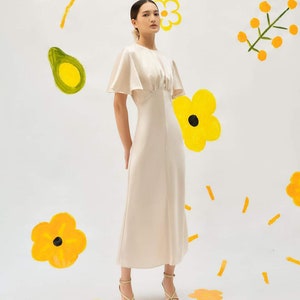 May include: A woman wearing a light beige midi dress with a round neckline and short, flared sleeves. She is standing on a white background with yellow painted flowers and avocado illustrations.